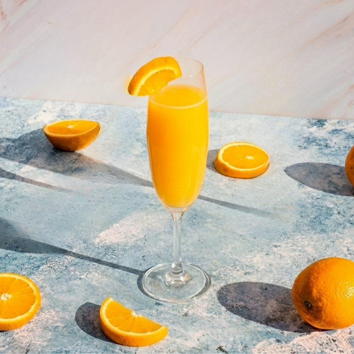 Un bodegón soleado con tres copas de flauta de champán llenas de un cóctel Mimosa de color naranja brillante, cada una decorada con una rodaja de naranja fresca en el borde. Las copas están dispuestas sobre una mesa con textura de piedra azul moteada, acompañadas de naranjas enteras y rodajas sueltas dispersas. El fondo es una pared de mármol veteado en tonos rosa pálido y blanco. La luz solar directa y dura proyecta sombras nítidas y largas de las copas y frutas hacia la izquierda.